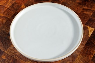 A plain, round, white ceramic plate is placed on a wooden surface with a checkered pattern made up of various shades of brown.