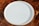 A plain, round, white ceramic plate is placed on a wooden surface with a checkered pattern made up of various shades of brown.