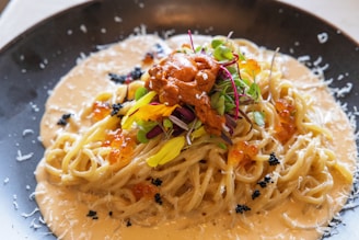 A creamy pasta dish topped with vibrant garnishes, including microgreens, fish roe, and grated cheese, served on a black plate. The sauce is rich and the pasta is elegantly twirled.
