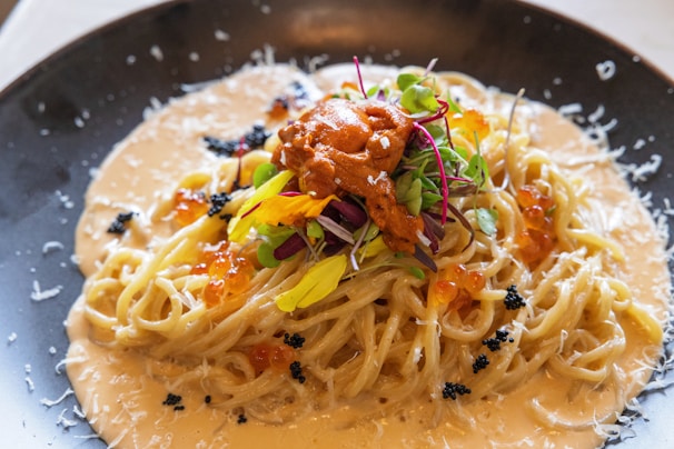 A creamy pasta dish topped with vibrant garnishes, including microgreens, fish roe, and grated cheese, served on a black plate. The sauce is rich and the pasta is elegantly twirled.