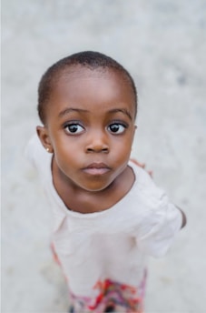 A young child with short hair is wearing a light-colored outfit. The child has a pensive expression and is looking upwards. The background is blurred, enhancing the focus on the child's face.
