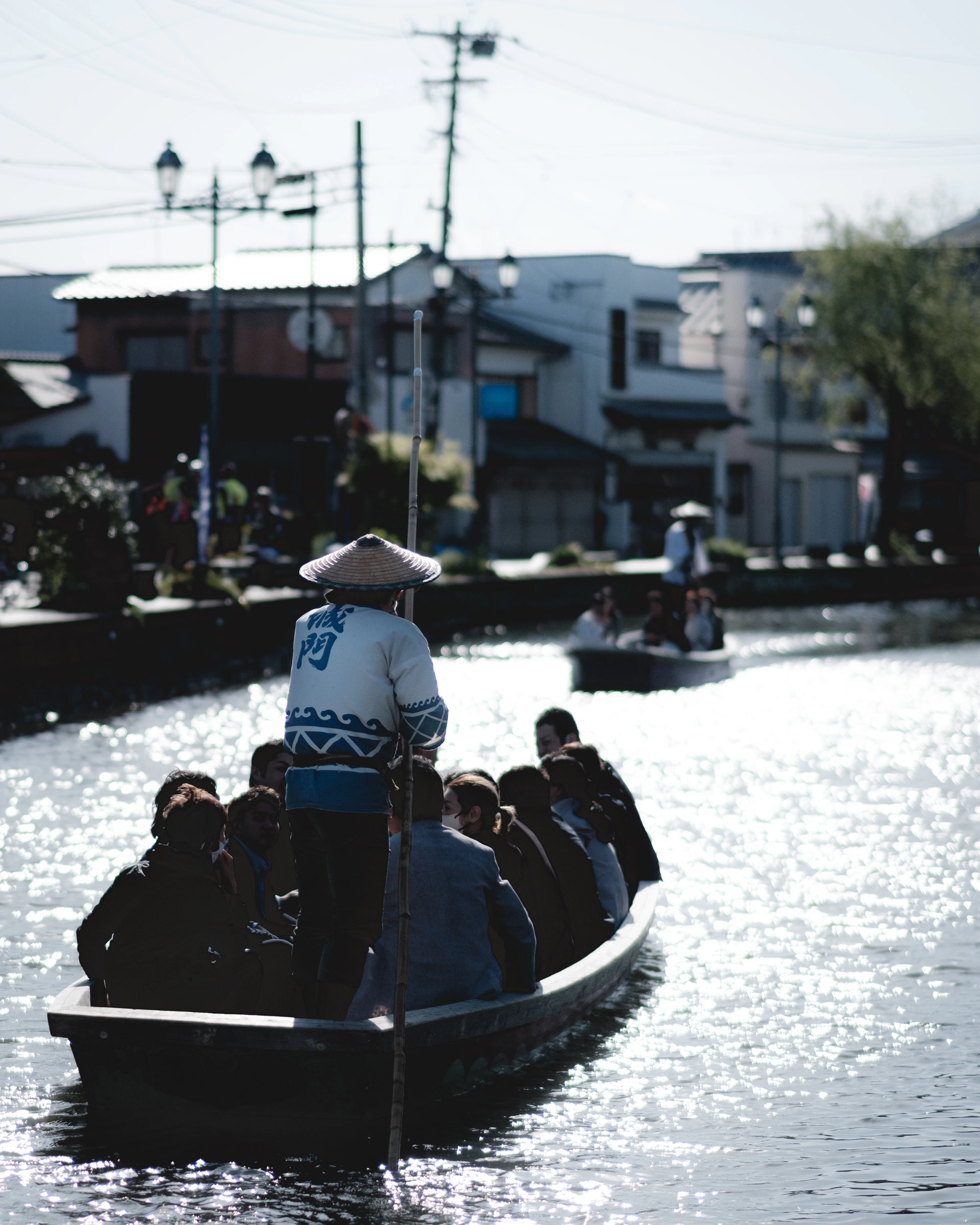 A boatman in a straw hat poles a long wooden punt crowded with passengers down a sunlit, sparkling canal.