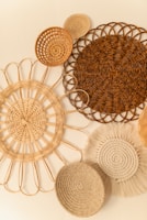 An arrangement of woven, circular rattan and wicker wall hangings in varying sizes and patterns. Each piece features a distinct design, ranging from tightly woven spirals to more open, looped structures. The assortment creates a textured and natural aesthetic against a light cream wall.