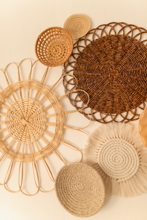 An arrangement of woven, circular rattan and wicker wall hangings in varying sizes and patterns. Each piece features a distinct design, ranging from tightly woven spirals to more open, looped structures. The assortment creates a textured and natural aesthetic against a light cream wall.