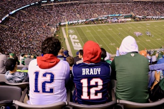 A crowded football stadium with fans sitting in the stands, wearing jerseys and hoodies. The field is visible in the background with teams lined up for a play. The audience is engaged, capturing the excitement of the game.