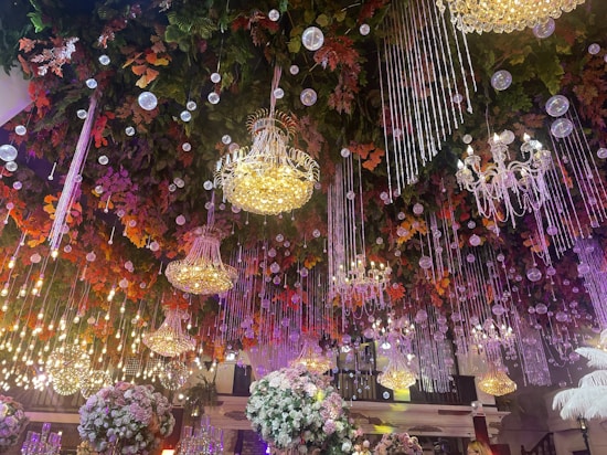 Luxurious chandeliers and crystal strings hang from a ceiling adorned with vibrant artificial foliage in shades of green, orange, and red. The decor includes clusters of white and pink flowers. Spherical light bulbs are intermixed, creating a twinkling effect.