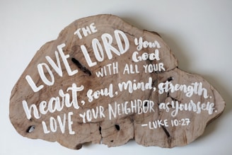 A piece of wood in an irregular shape with a religious text painted in white. The text reads: 'The LOVE LORD your God with all your heart, soul, mind, strength LOVE your neighbor as yourself. -Luke 10:27'. The wood grain is visible and has a natural, rustic appearance.