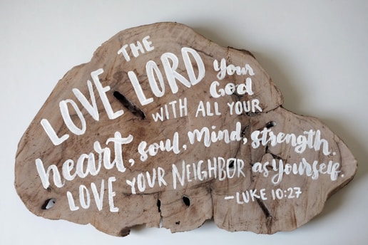 A piece of wood in an irregular shape with a religious text painted in white. The text reads: 'The LOVE LORD your God with all your heart, soul, mind, strength LOVE your neighbor as yourself. -Luke 10:27'. The wood grain is visible and has a natural, rustic appearance.