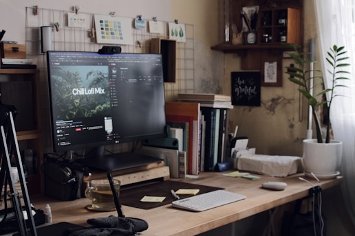 A cozy and cluttered home office desk setup with a large computer monitor displaying a video titled 'Chill Lofi Mix'. The desk is adorned with books, a keyboard, a mouse, sticky notes, and a small plant. To the right, a window with sheer curtains allows natural light to filter in. The wall has decorative items like inspirational quotes and a small shelf with various objects.