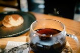 A glass mug filled with dark coffee sits on a wooden surface. In the background, there is a blurred view of a pancake on a black plate. A partially visible card labeled 'ETHIO' is seen beneath the mug.