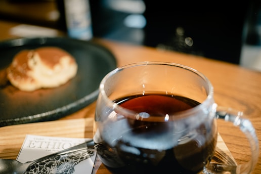 A glass mug filled with dark coffee sits on a wooden surface. In the background, there is a blurred view of a pancake on a black plate. A partially visible card labeled 'ETHIO' is seen beneath the mug.