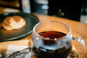 A glass mug filled with dark coffee sits on a wooden surface. In the background, there is a blurred view of a pancake on a black plate. A partially visible card labeled 'ETHIO' is seen beneath the mug.