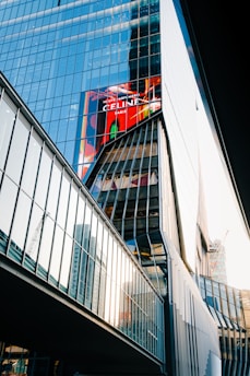 A modern glass building with reflective windows that capture the surrounding structures and sky. The architecture is angular and features a prominent digital billboard displaying a colorful advertisement. Adjacent structures are visible, and there is construction equipment in the background.