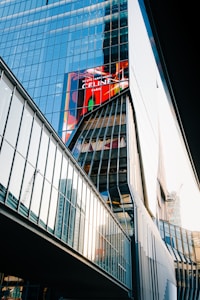 A modern glass building with reflective windows that capture the surrounding structures and sky. The architecture is angular and features a prominent digital billboard displaying a colorful advertisement. Adjacent structures are visible, and there is construction equipment in the background.