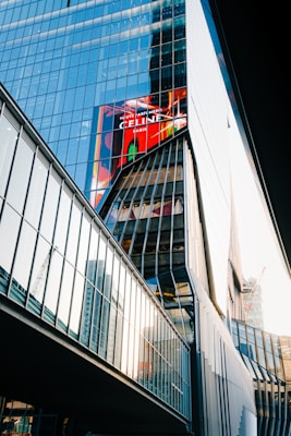 A modern glass building with reflective windows that capture the surrounding structures and sky. The architecture is angular and features a prominent digital billboard displaying a colorful advertisement. Adjacent structures are visible, and there is construction equipment in the background.