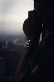 A person is looking through a viewfinder or a camera, with a visible wristwatch on their arm. The setting appears dimly lit, suggesting a contemplative or introspective moment. The background is blurred, indicating a focus on the subject.