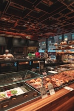 A bakery counter displays a variety of fresh pastries and baked goods under a glass case. Behind the counter, there are ovens and kitchen equipment, with bakers attending to their tasks. The setting has a warm, inviting ambiance with wooden and metallic elements, and a ceiling with geometric patterns. Large windows offer a glimpse of the urban street outside.