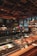 A bakery counter displays a variety of fresh pastries and baked goods under a glass case. Behind the counter, there are ovens and kitchen equipment, with bakers attending to their tasks. The setting has a warm, inviting ambiance with wooden and metallic elements, and a ceiling with geometric patterns. Large windows offer a glimpse of the urban street outside.
