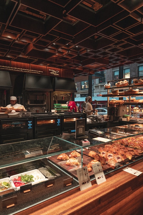 A bakery counter displays a variety of fresh pastries and baked goods under a glass case. Behind the counter, there are ovens and kitchen equipment, with bakers attending to their tasks. The setting has a warm, inviting ambiance with wooden and metallic elements, and a ceiling with geometric patterns. Large windows offer a glimpse of the urban street outside.
