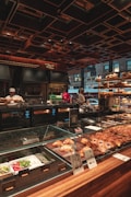 A bakery counter displays a variety of fresh pastries and baked goods under a glass case. Behind the counter, there are ovens and kitchen equipment, with bakers attending to their tasks. The setting has a warm, inviting ambiance with wooden and metallic elements, and a ceiling with geometric patterns. Large windows offer a glimpse of the urban street outside.