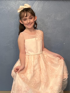 A young girl with brown hair is smiling while wearing a light pink dress with floral embroidery and a pearl belt, standing against a textured blue wall. She has a large bow in her hair and is holding the sides of her dress.