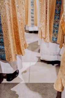 Golden ornate fabric with intricate patterns draped over white garments, worn by several individuals whose lower legs and black shoes are visible. The fabric includes a central strip of detailed blue embroidery.