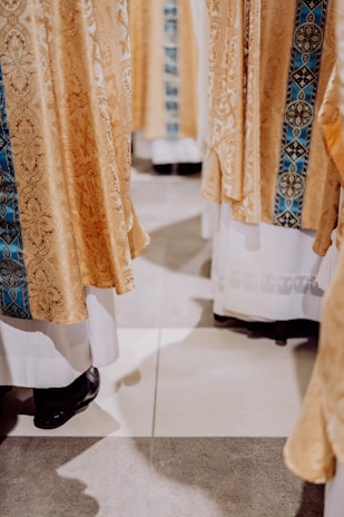 Golden ornate fabric with intricate patterns draped over white garments, worn by several individuals whose lower legs and black shoes are visible. The fabric includes a central strip of detailed blue embroidery.