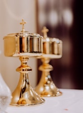 Two ornate, golden chalices with cross engravings on their lids are placed on a white surface. They have a polished finish and reflect the surrounding light, creating a warm glow. The background is softly blurred, emphasizing the focus on the chalices.