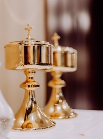 Two ornate, golden chalices with cross engravings on their lids are placed on a white surface. They have a polished finish and reflect the surrounding light, creating a warm glow. The background is softly blurred, emphasizing the focus on the chalices.