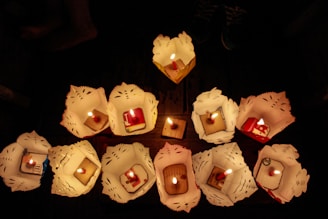 A group of intricately folded paper lanterns with lit candles inside are arranged on a wooden surface in a dimly lit setting. The lanterns feature decorative cut-outs and each holds a brightly burning candle in the center.