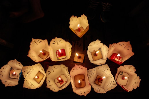 A group of intricately folded paper lanterns with lit candles inside are arranged on a wooden surface in a dimly lit setting. The lanterns feature decorative cut-outs and each holds a brightly burning candle in the center.