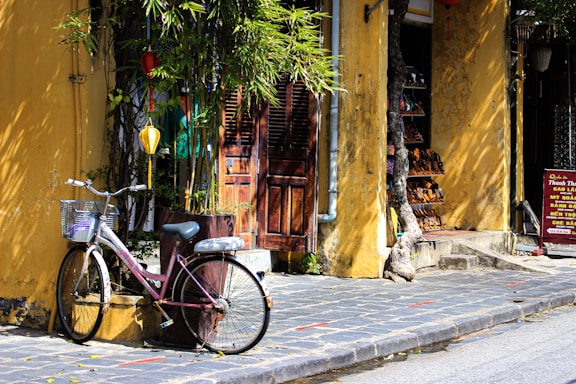 A vintage bicycle with a wire basket is parked against a yellow, textured wall with a wooden door. Overhead lanterns hang from a lush green tree, providing a quaint, serene atmosphere. A small shop with rows of shoes displayed on shelves is partially visible, while a sign with Vietnamese text stands beside the entrance.