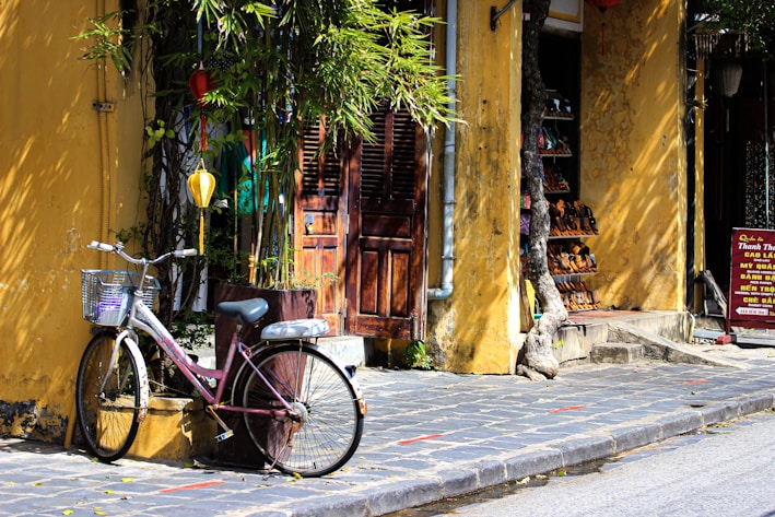A vintage bicycle with a wire basket is parked against a yellow, textured wall with a wooden door. Overhead lanterns hang from a lush green tree, providing a quaint, serene atmosphere. A small shop with rows of shoes displayed on shelves is partially visible, while a sign with Vietnamese text stands beside the entrance.