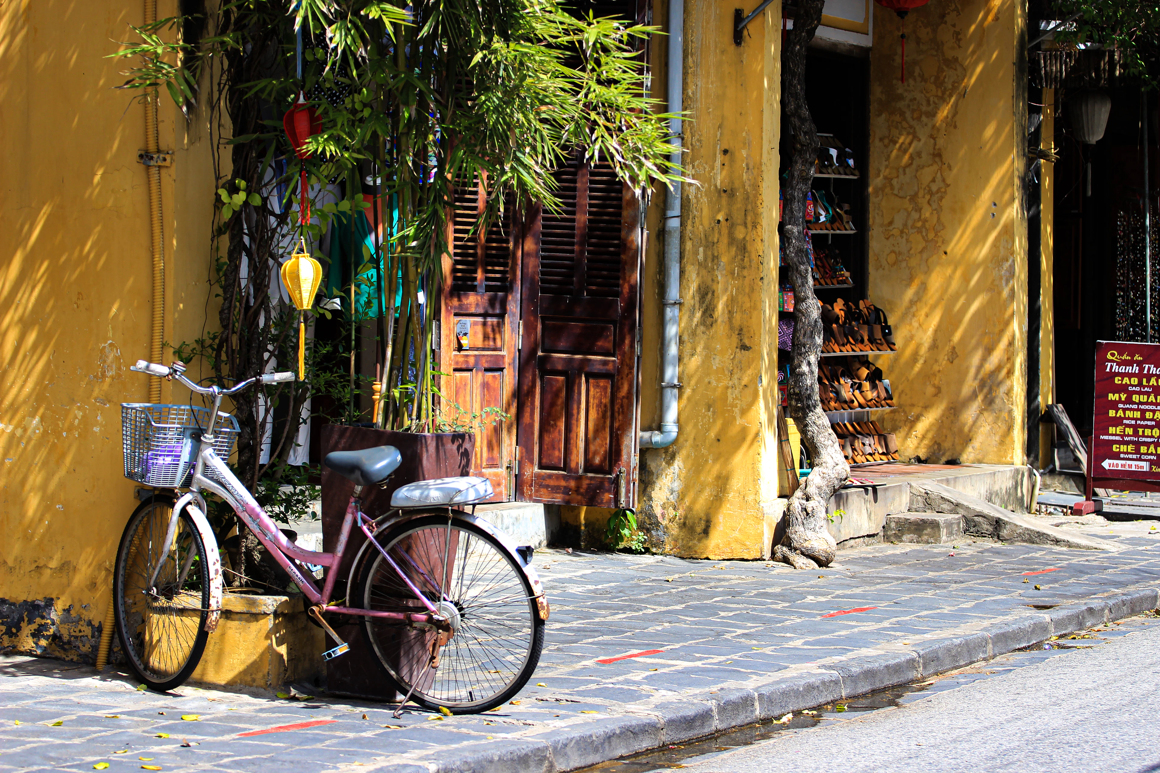 A bicycle with a wicker basket leaning against the weathered ochre wall of a Hoi An Ancient Town lane, beside carved wooden shutters, a hanging lantern and a souvenir stall