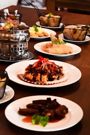 A dining table is set with various Asian dishes served on white plates. A notable dish features roasted meat covered in a dark sauce, garnished with red chili slices and green herbs. There is a metal hot pot containing soup or broth, baskets with side items like spring rolls, and plates of sliced meat and fried appetizers.