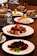 A dining table is set with various Asian dishes served on white plates. A notable dish features roasted meat covered in a dark sauce, garnished with red chili slices and green herbs. There is a metal hot pot containing soup or broth, baskets with side items like spring rolls, and plates of sliced meat and fried appetizers.