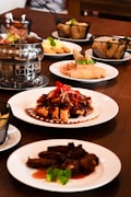 A dining table is set with various Asian dishes served on white plates. A notable dish features roasted meat covered in a dark sauce, garnished with red chili slices and green herbs. There is a metal hot pot containing soup or broth, baskets with side items like spring rolls, and plates of sliced meat and fried appetizers.