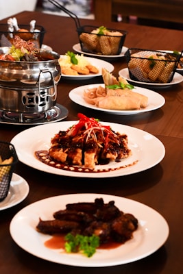 A dining table is set with various Asian dishes served on white plates. A notable dish features roasted meat covered in a dark sauce, garnished with red chili slices and green herbs. There is a metal hot pot containing soup or broth, baskets with side items like spring rolls, and plates of sliced meat and fried appetizers.