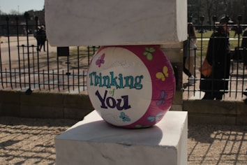 A colorful balloon with the words 'Thinking of You' written on it is placed between two stone blocks. The balloon features butterfly designs and is predominantly pink and white. In the background, there is a metal fence and some people walking, with a road and trees visible in the distance.