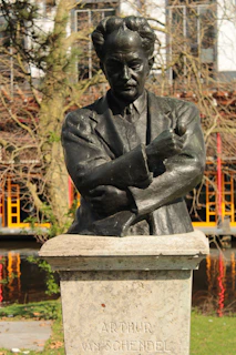A bronze statue of a man with an assertive posture, featuring crossed arms and a focused expression. The statue is mounted on a stone pedestal with the name 'Arthur van Schendel' engraved on it. In the background, there are trees with bare branches and a building with scaffolding, indicating construction work.