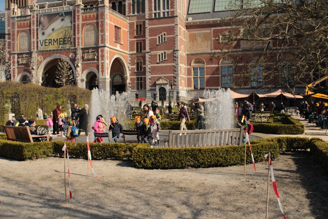 A group of children wearing brightly colored hats walk near a fountain in a courtyard with neatly trimmed hedges. A large brick building with arches and an advertisement for a Vermeer exhibition is in the background. Adults are scattered around, some watching the children and others sitting on benches. The scene is lively and bathed in sunlight.