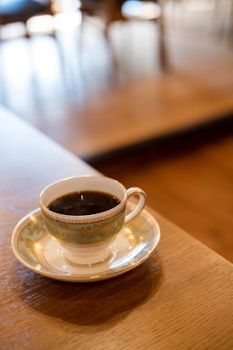 A delicate porcelain teacup filled with black coffee rests on a matching saucer. The set features ornate gold decoration against a subtle blue-green background, all placed on a polished wooden table. The background is softly blurred, indicating an indoor setting, possibly a cozy cafe.