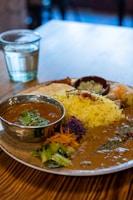 A plate of assorted Indian food with a metal bowl of curry garnished with herbs. The plate includes yellow rice, purple cabbage, carrots, and various vegetables. A glass of water is placed next to the plate on a wooden table.