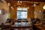 A cozy cafe interior with wooden furniture and warm lighting. The space is decorated with framed art on the walls and books on shelves. Several tables and chairs are arranged neatly, and there's a large window at the far end allowing natural light to come in.