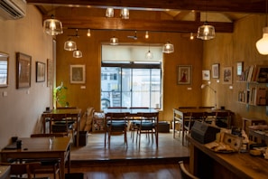 A cozy cafe interior with wooden furniture and warm lighting. The space is decorated with framed art on the walls and books on shelves. Several tables and chairs are arranged neatly, and there's a large window at the far end allowing natural light to come in.