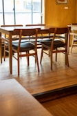 A wooden table surrounded by several chairs with black cushions is placed on a wooden floor in a well-lit room. A window in the background allows natural light to illuminate the space, highlighting the texture and warmth of the wood. The ambiance appears simple and cozy, with minimal decoration visible.