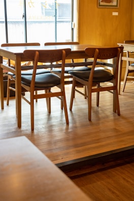 A wooden table surrounded by several chairs with black cushions is placed on a wooden floor in a well-lit room. A window in the background allows natural light to illuminate the space, highlighting the texture and warmth of the wood. The ambiance appears simple and cozy, with minimal decoration visible.