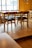 A wooden table surrounded by several chairs with black cushions is placed on a wooden floor in a well-lit room. A window in the background allows natural light to illuminate the space, highlighting the texture and warmth of the wood. The ambiance appears simple and cozy, with minimal decoration visible.