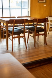 A wooden table surrounded by several chairs with black cushions is placed on a wooden floor in a well-lit room. A window in the background allows natural light to illuminate the space, highlighting the texture and warmth of the wood. The ambiance appears simple and cozy, with minimal decoration visible.