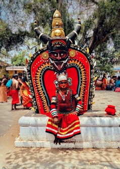 A person wearing a traditional red and black costume with intricate patterns and a headdress sits in front of a large, ornate backdrop with a demonic face and horns. The backdrop is decorated with bold colors and intricate designs. Surrounding the person, other individuals are dressed in similar cultural attire. The setting appears to be outdoors with trees and other people visible in the background.